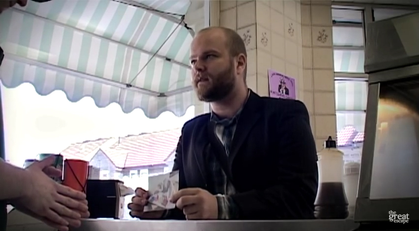 A still from The Terror of the Killer Carnivorous Coat. The lead character Gabriel Cushing stands at a chip shop counter, enquiring about the photo in his hand.