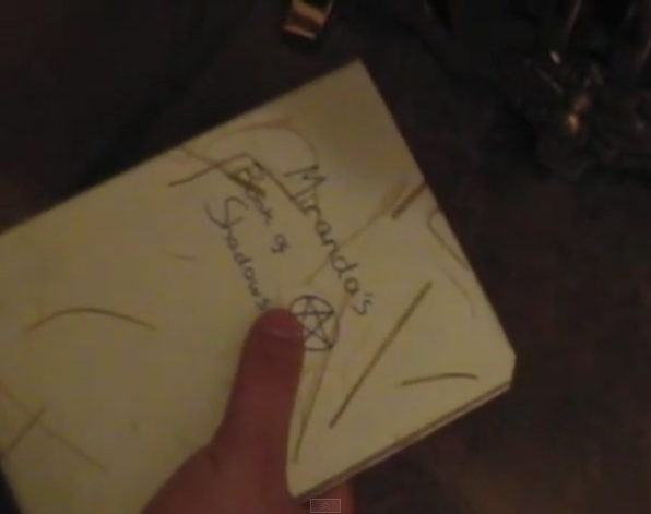 A still from Threefold. A white book with the words Miranda's Book of Shadows written on the front in pen, together with a pentagram doodle.